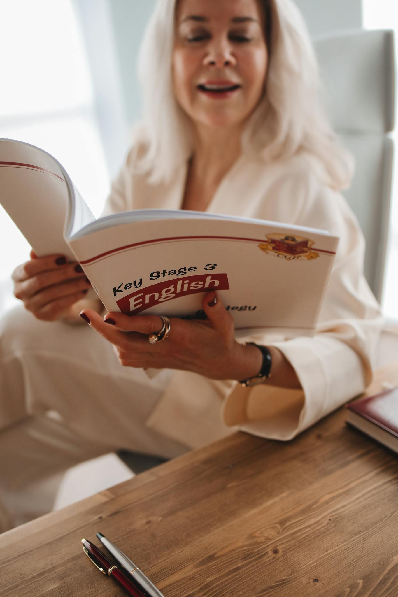 Adult woman reading a Key Stage 3 English book, seated at a wooden table.
